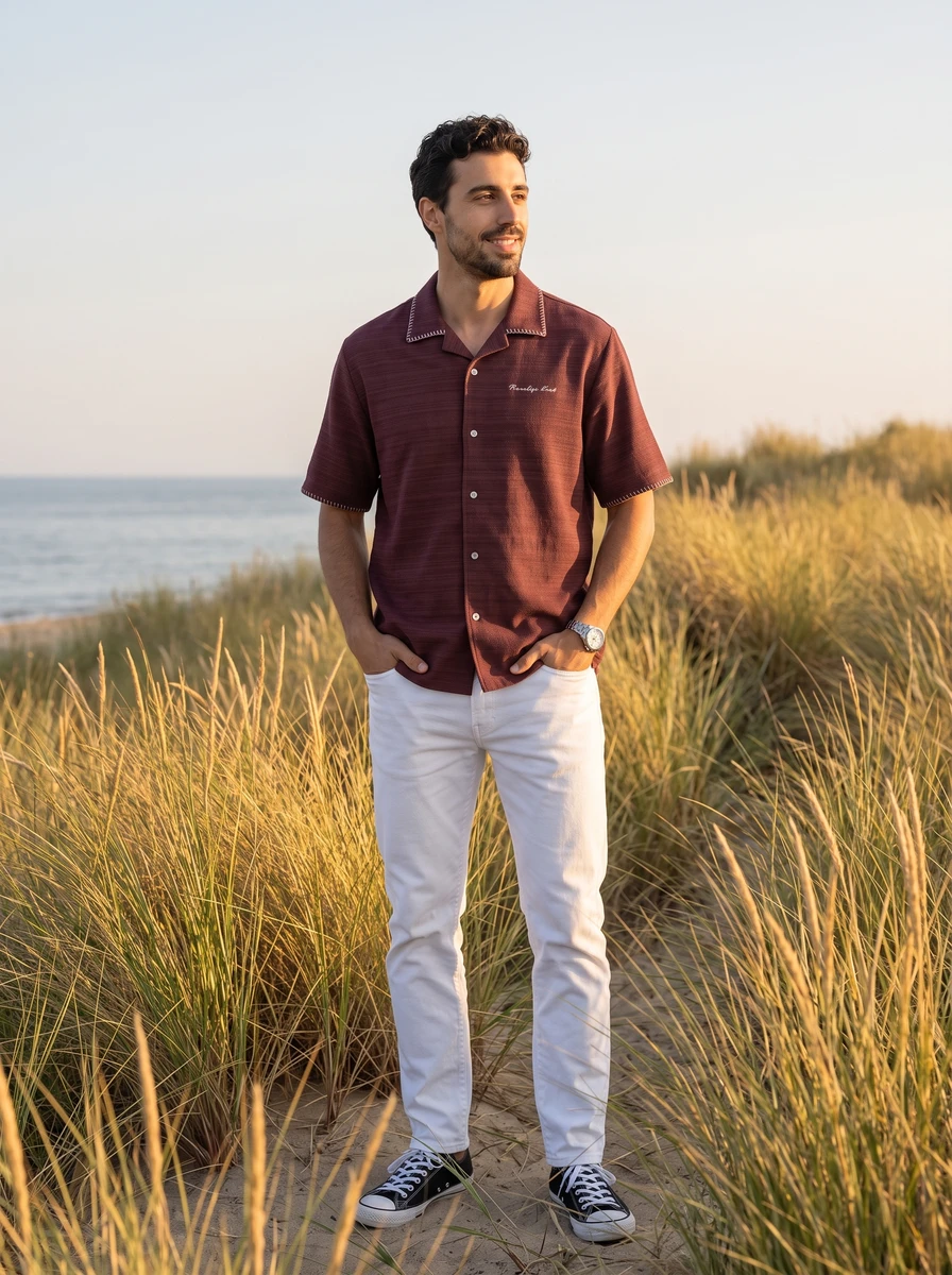 A man wearing a maroon short-sleeve camp shirt and white jeans standing on a grassy sand dune near the ocean.