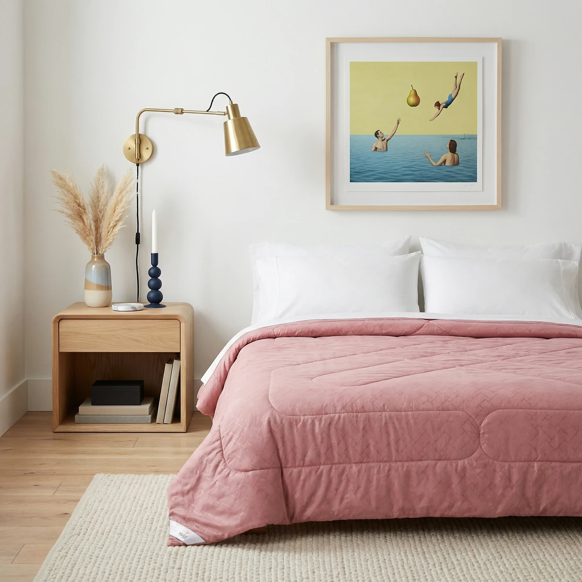 A pink textured comforter on a bed with white pillows in a brightly lit bedroom with a wooden nightstand.