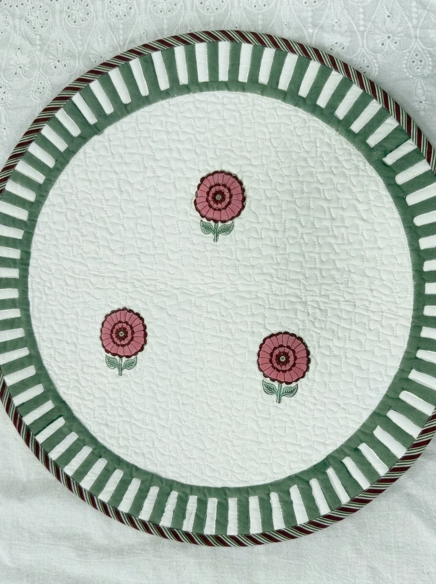 Round quilted table mat in white with three pink flower motifs and a green striped border.