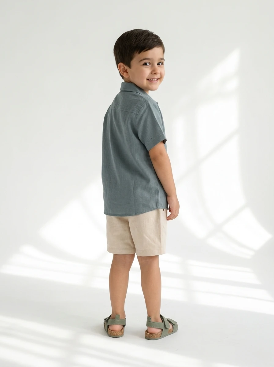 Young boy wearing a blue short-sleeved button-down shirt and beige shorts, smiling while looking over his shoulder.
