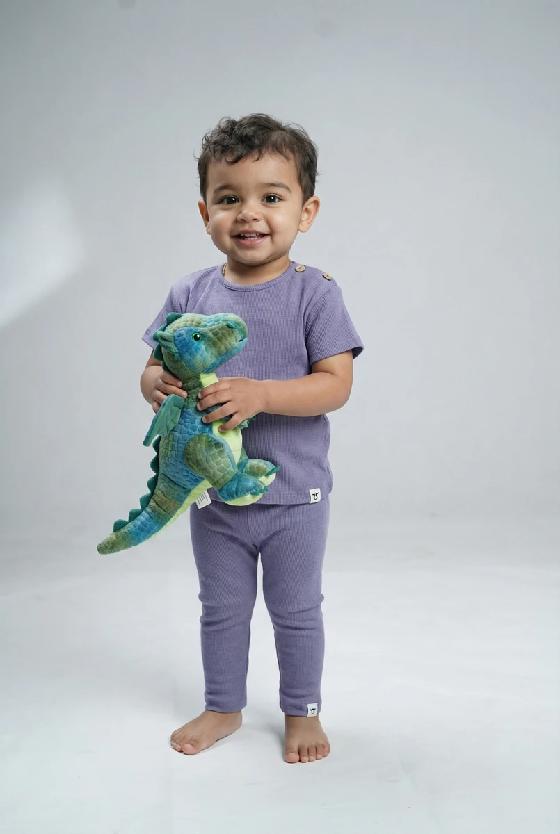 Toddler smiling while holding a stuffed green dinosaur, wearing a purple ribbed two-piece apparel set against a light background.