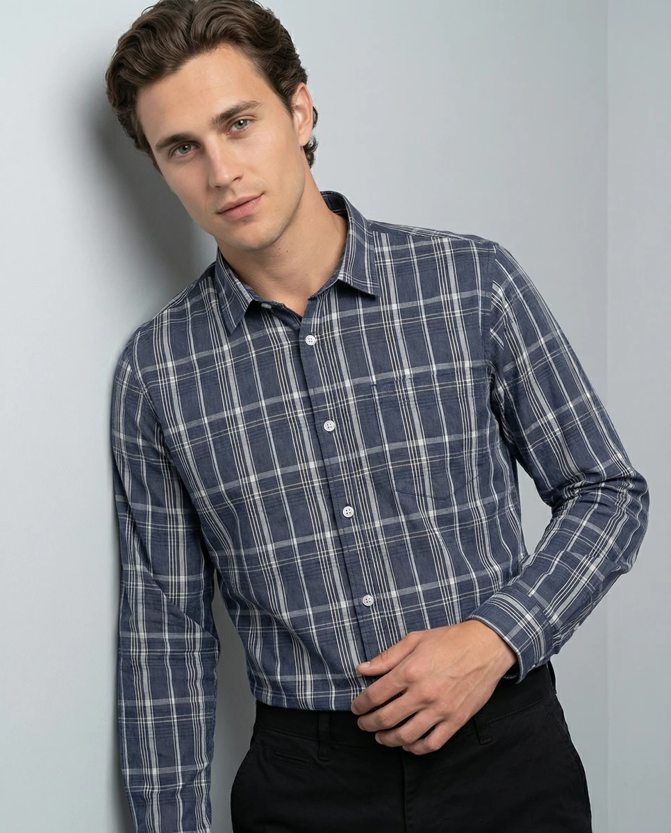 Man wearing a blue and white plaid long-sleeve button-down shirt leaning against a grey wall