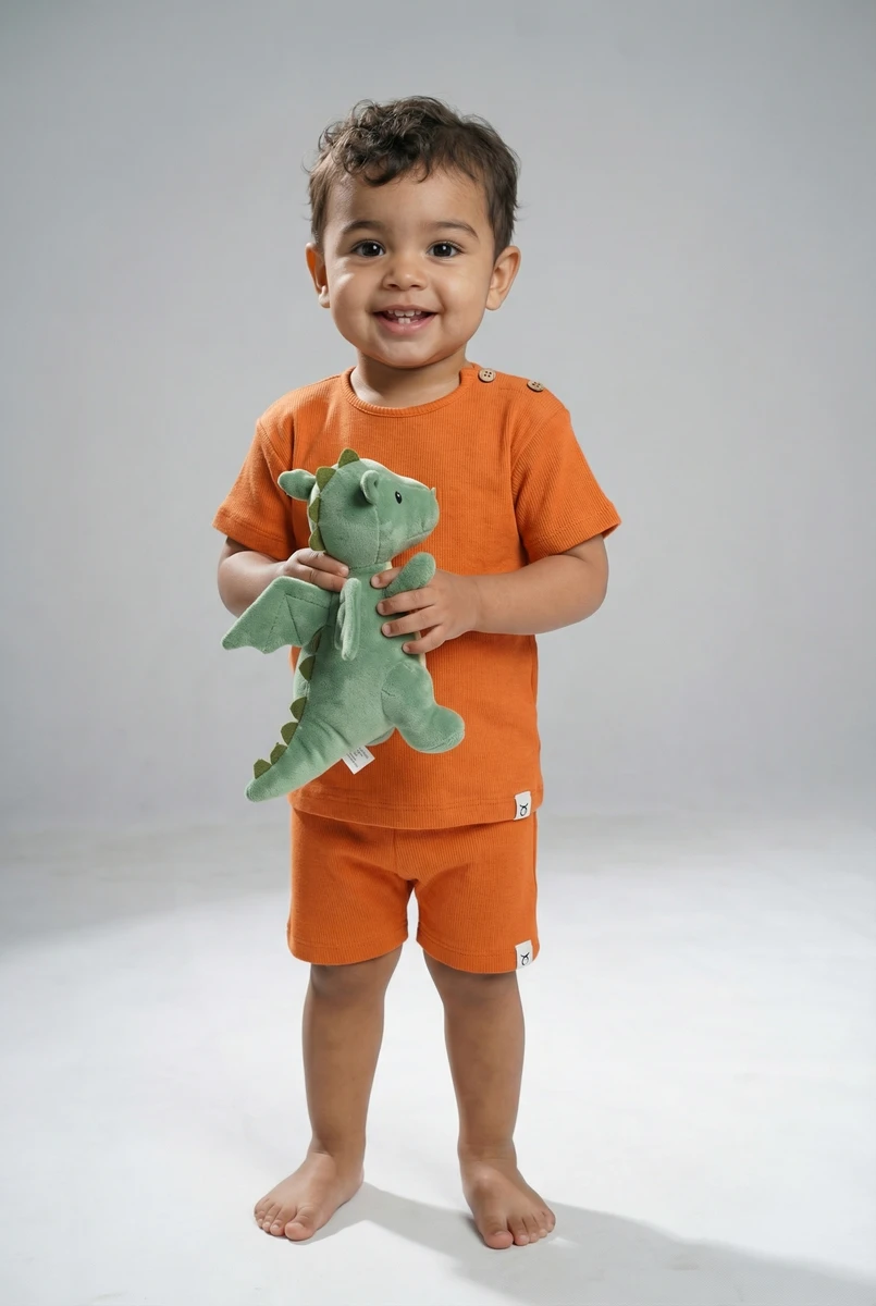 Toddler wearing an orange ribbed short-sleeve shirt and matching shorts, smiling while holding a green plush dragon toy.