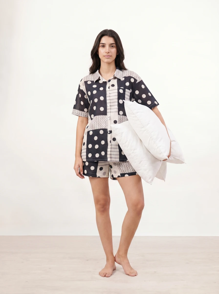 Woman standing in black and white polka dot pajama set holding white pillows against a plain background