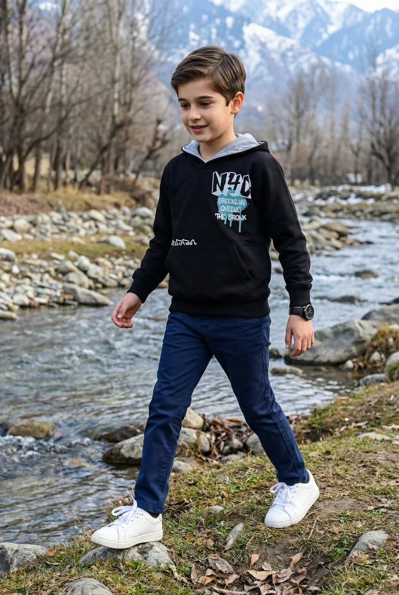 Young boy walking by a mountain stream wearing a black hoodie with graphic text, blue pants, and white sneakers.