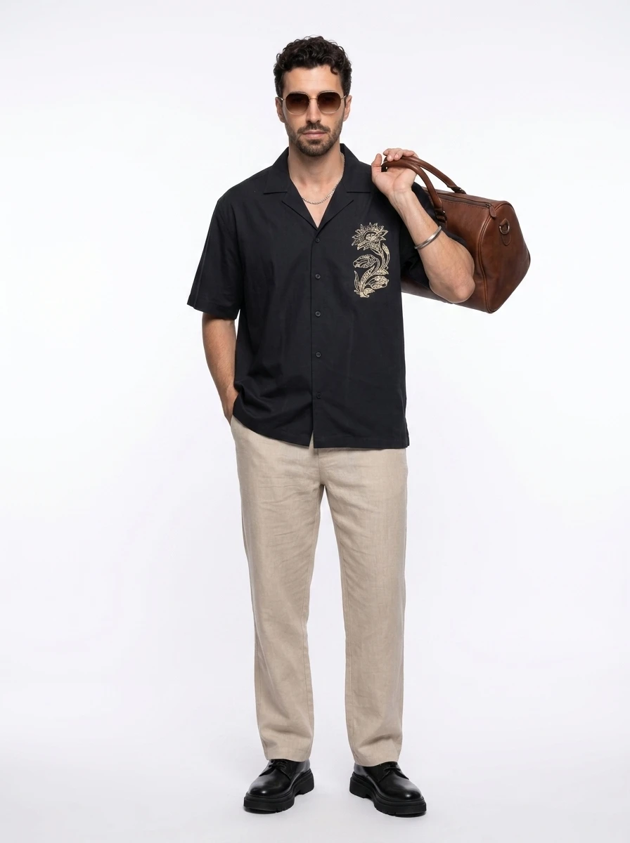 Man wears a black camp collar shirt with gold floral embroidery, beige linen trousers, and carries a brown leather bag.
