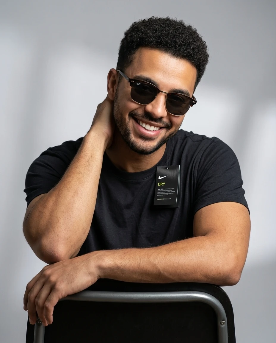 Smiling man in a black crew neck t-shirt wearing sunglasses, leaning on a chair against a gray studio backdrop.