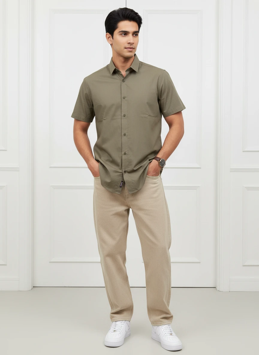 A man wearing an olive green short-sleeve button-down shirt and khaki pants, standing indoors against a white paneled wall.