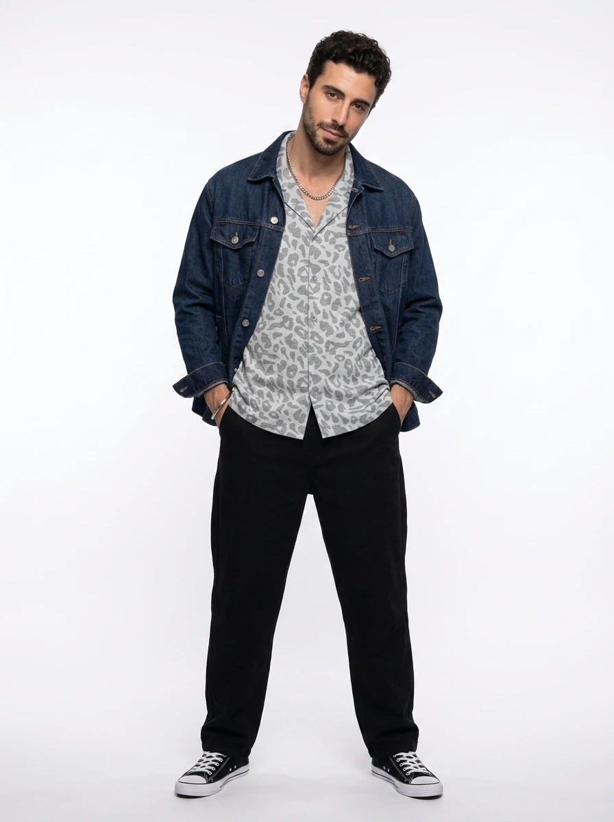 Man standing in dark denim jacket, grey animal print camp shirt, and black trousers against white backdrop.
