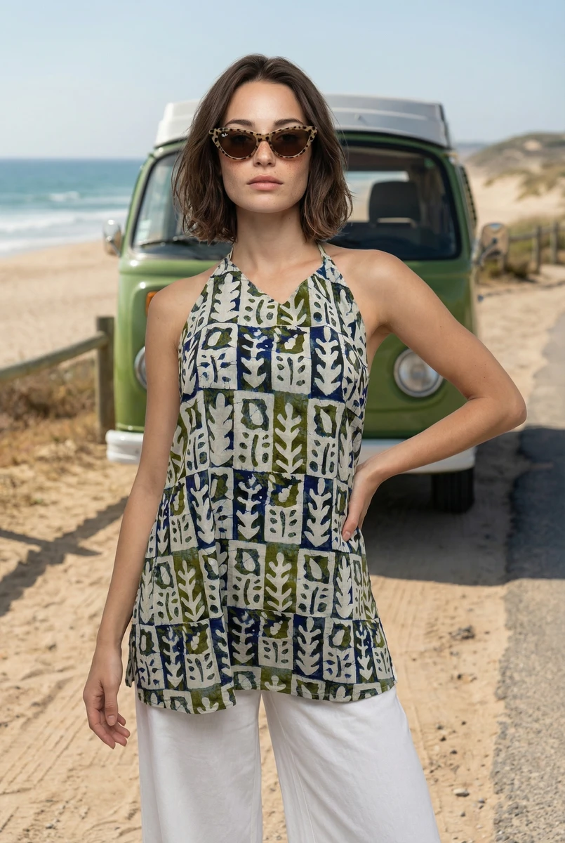 Woman in sleeveless blue and green patterned top and white pants poses on a beach by a green vintage van.