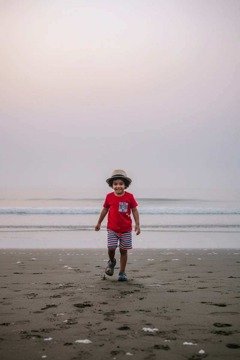 Young boy wearing a red t-shirt and striped shorts walks along a sandy beach under an overcast sky.