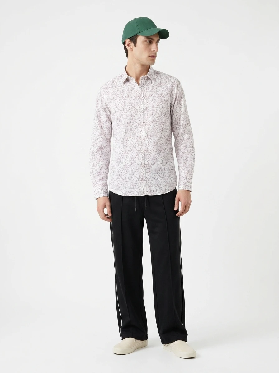 Man standing against a white backdrop wearing a patterned long-sleeve shirt, black trousers, green cap, and light slip-on shoes.