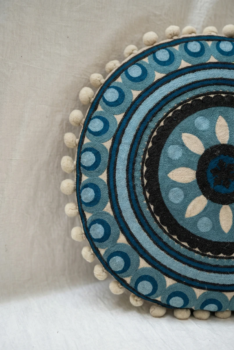 Sanskruti round blue cushion featuring concentric embroidered patterns and a pom pom trim.