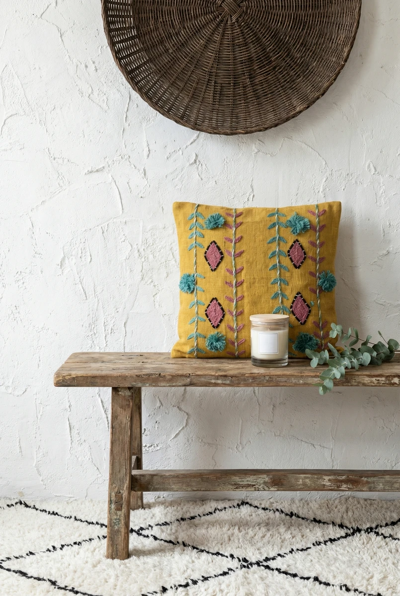 Yellow embroidered cushion with pink diamond patterns and teal pom-poms on a rustic wooden bench.