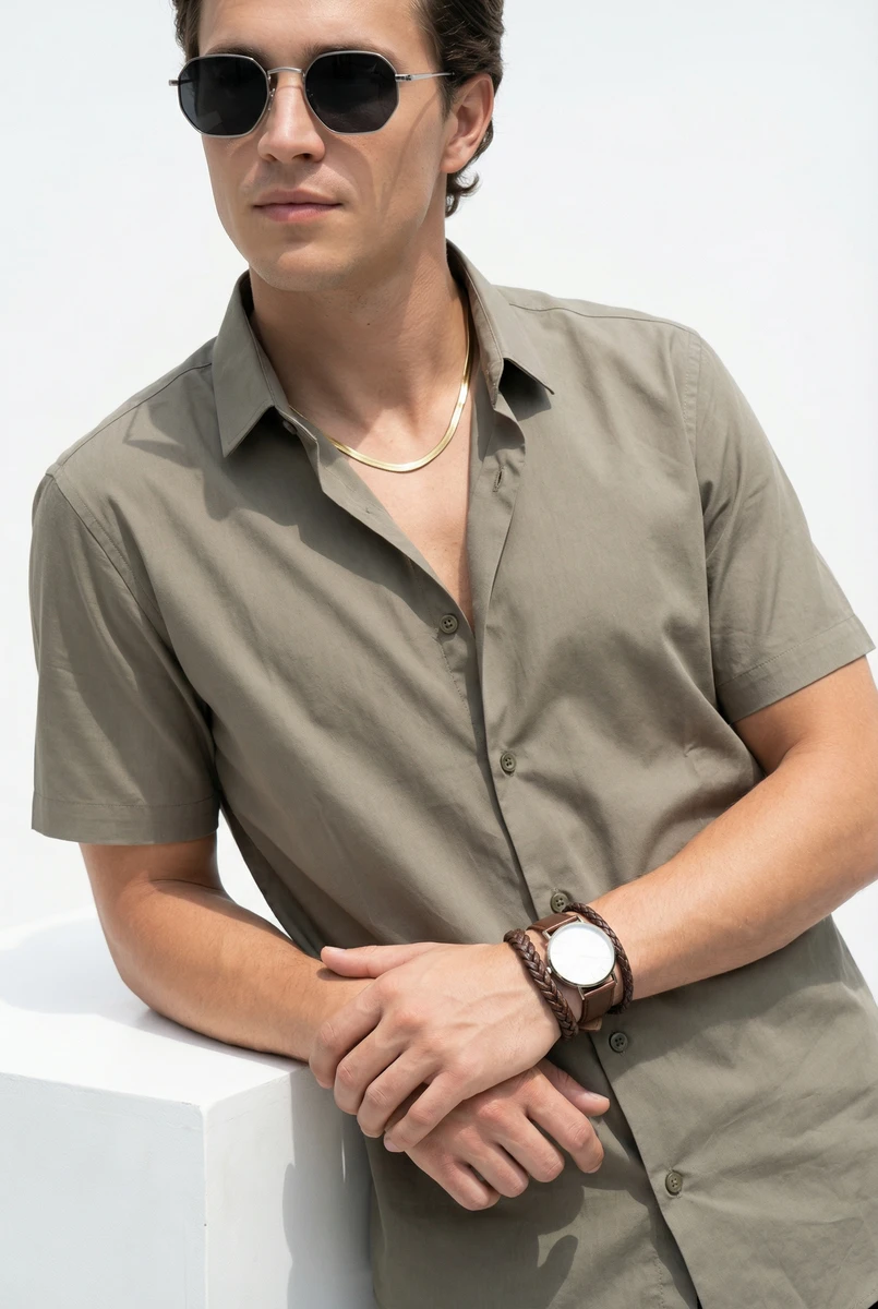A man wearing an olive green short-sleeve button-down shirt, sunglasses, and a watch against a white backdrop.