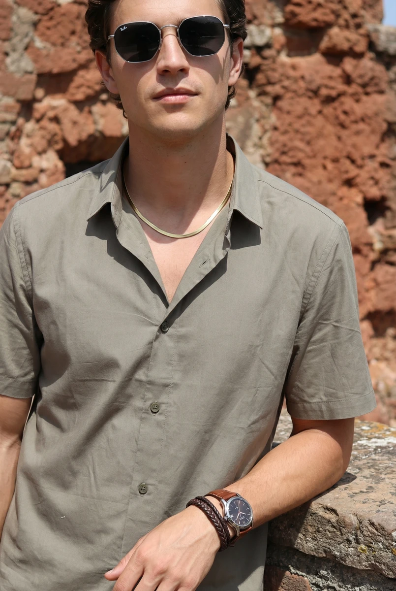 Man wearing a grey short-sleeve button-down shirt and sunglasses outdoors against a rustic stone wall background.