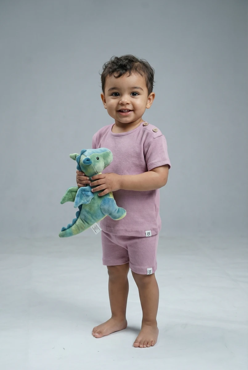 A smiling toddler wears a dusty pink ribbed short-sleeve set with button details while holding a green plush dragon toy.