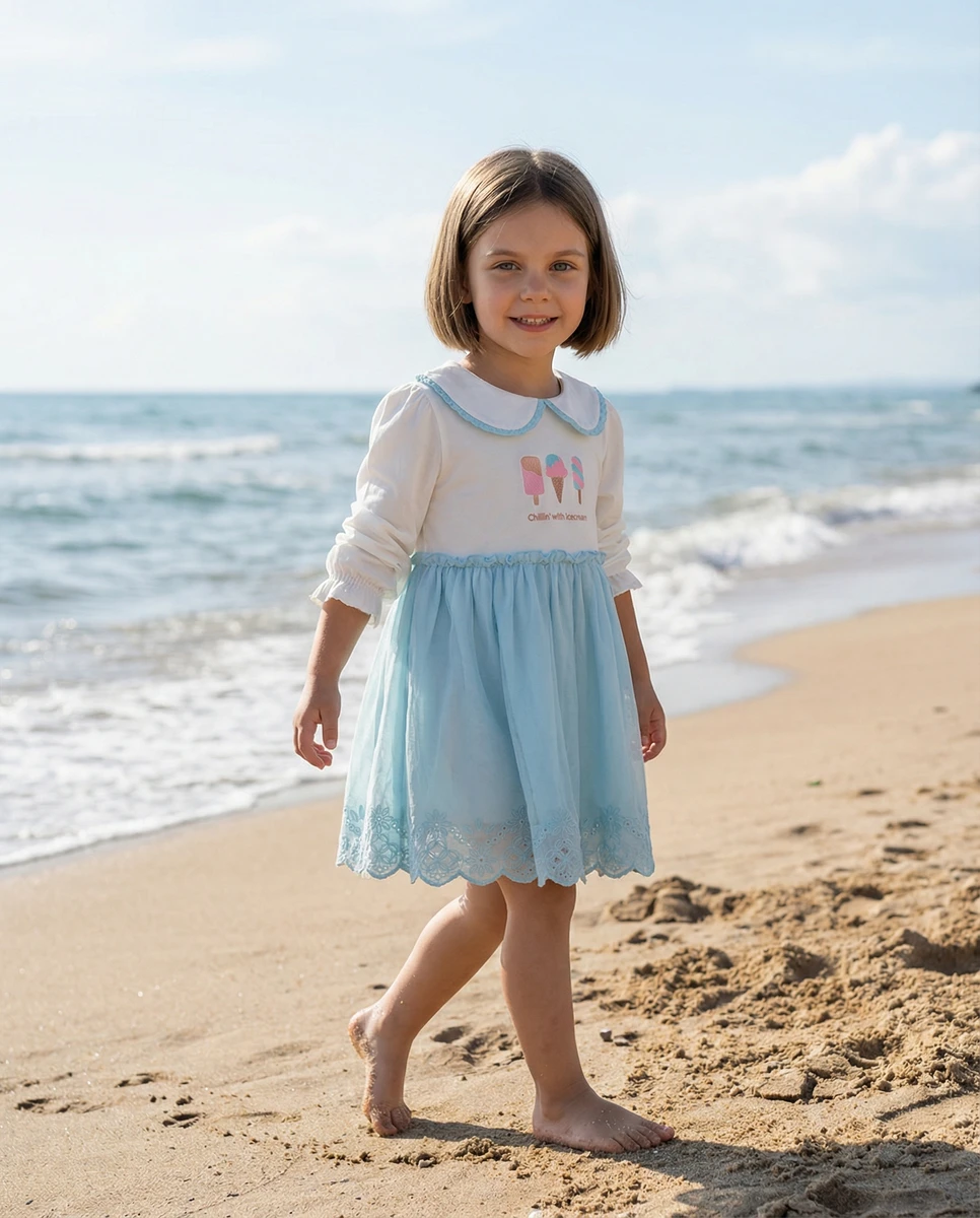 Young girl wearing a long sleeve white and blue dress with ice cream print walking on a sandy beach.