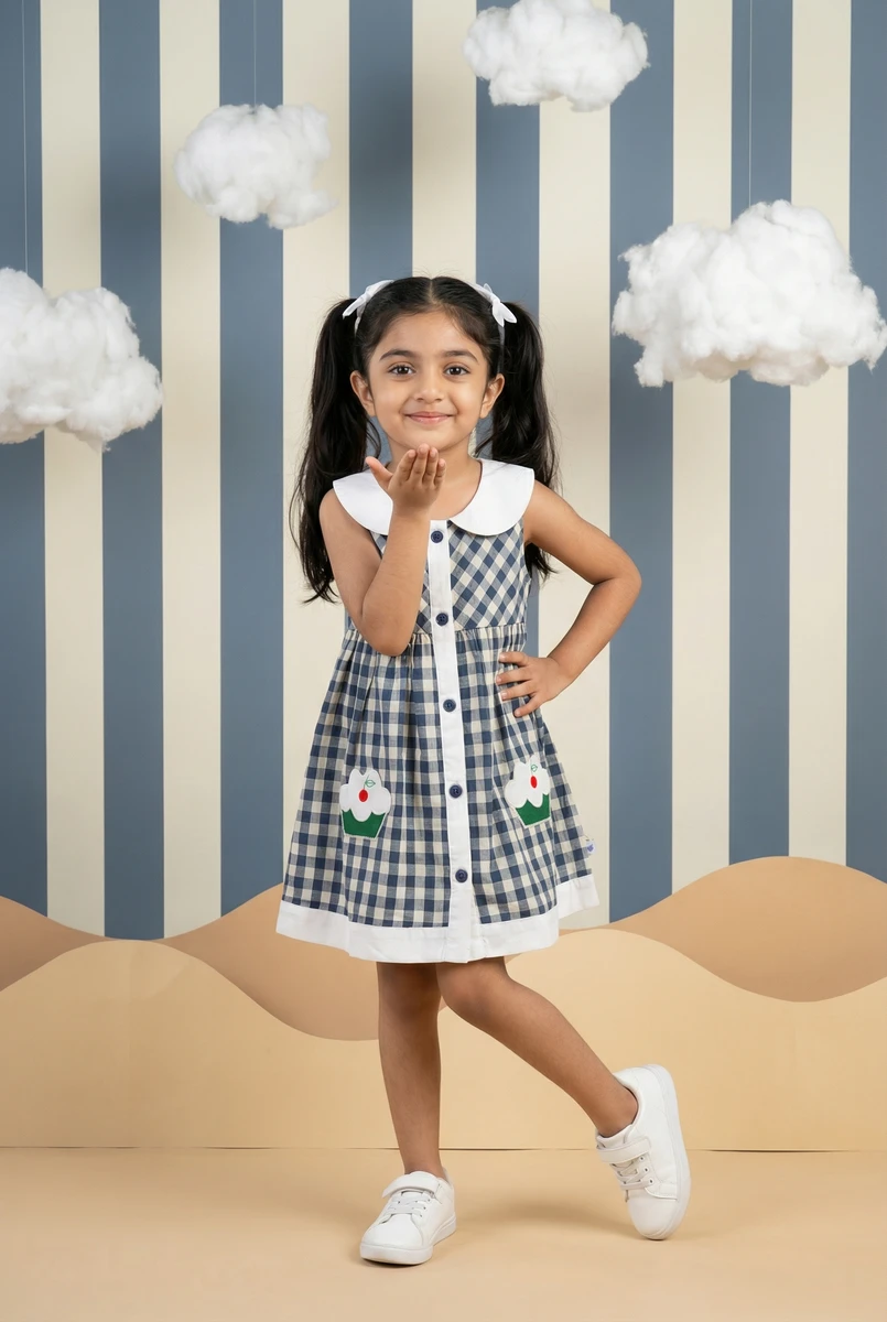 Young girl in a blue and white gingham dress with cupcake pockets posing against a striped wall with hanging clouds.