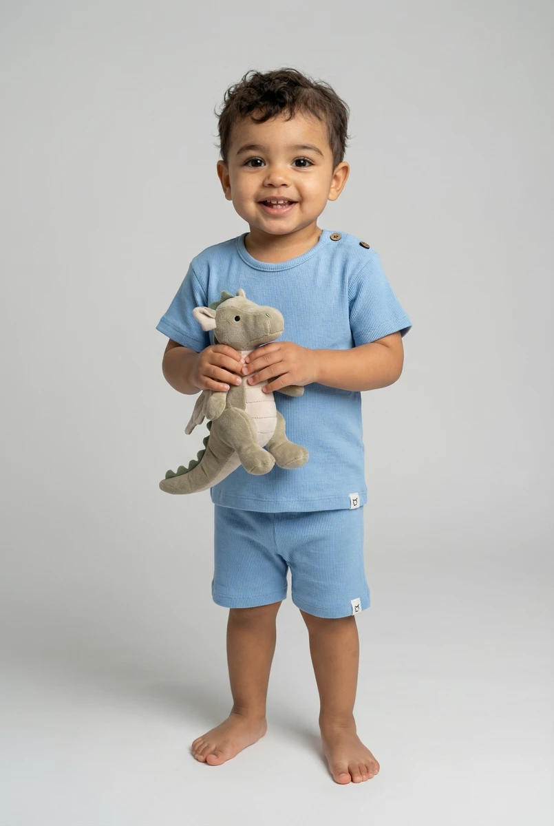 Toddler boy standing in blue ribbed cotton short set holding a stuffed dinosaur.