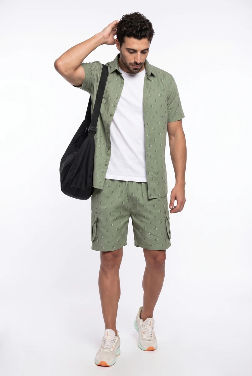 Man wearing a sage green patterned button-down shirt and cargo shorts, layered over a white t-shirt, carrying a black bag.