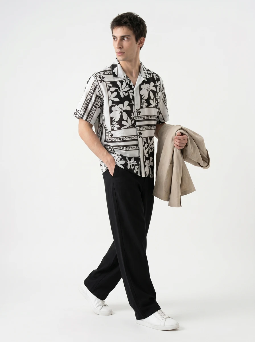 A man wearing a black and white floral short-sleeve shirt, black trousers, and white sneakers, holding a beige jacket.