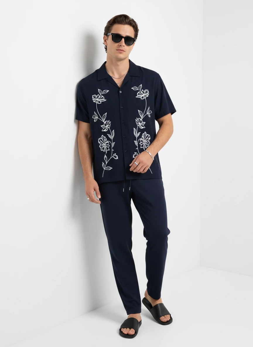 Man wearing a navy short-sleeve shirt with white floral embroidery and matching trousers, standing against a white wall.