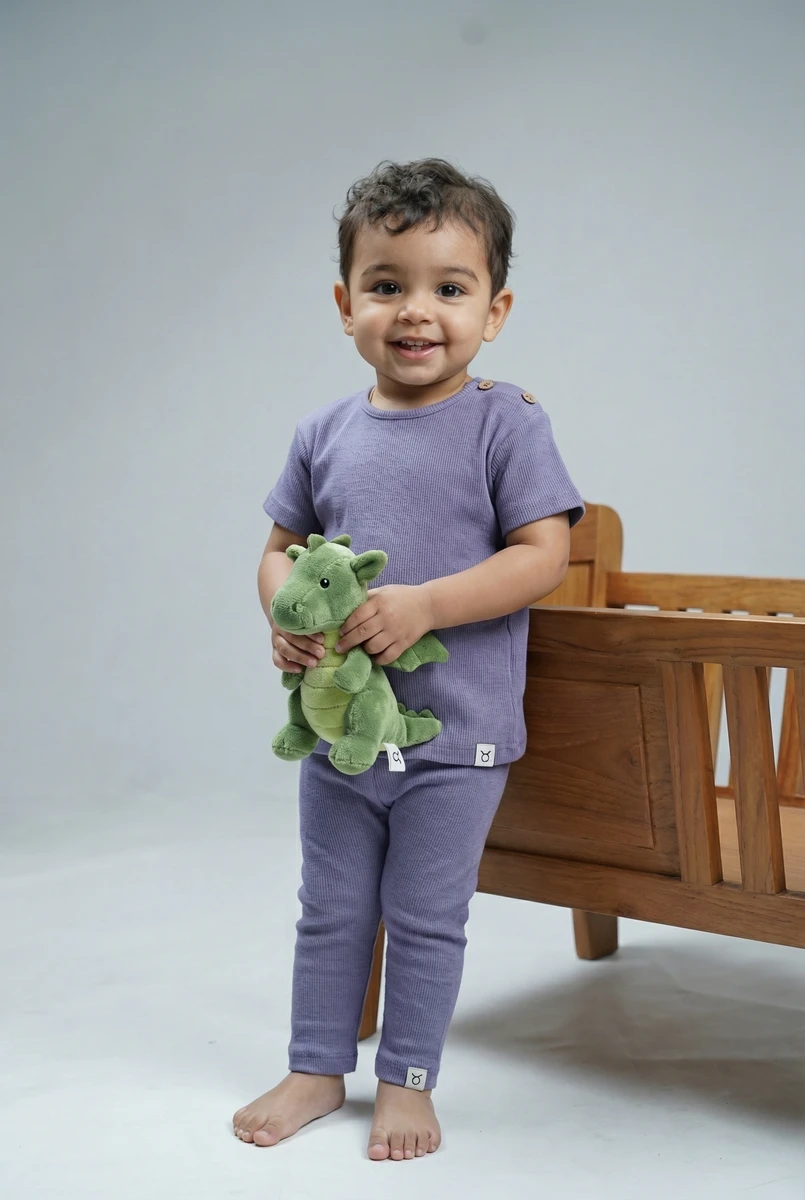 Toddler smiling while wearing a lavender ribbed short-sleeve top and matching pants, holding a green stuffed dragon.