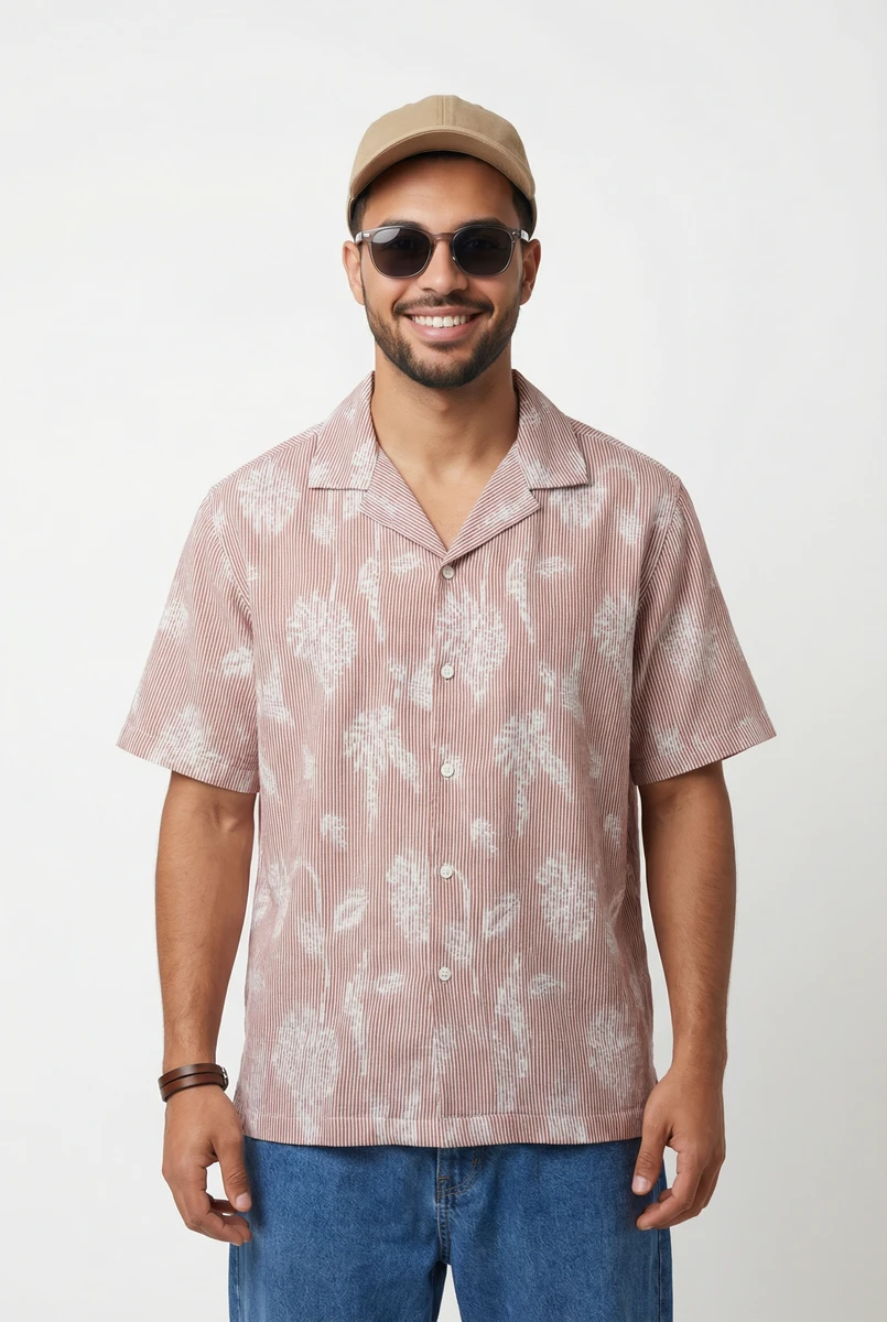 Man wearing a pink and white patterned short sleeve shirt, blue jeans, sunglasses, and a tan cap.