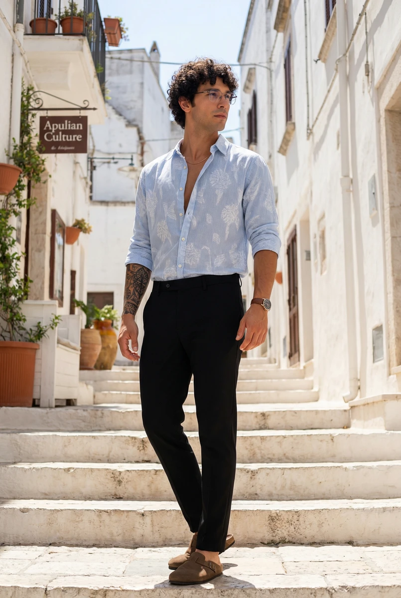 Man wearing a blue and white patterned shirt and black trousers, posing on stone steps outdoors.