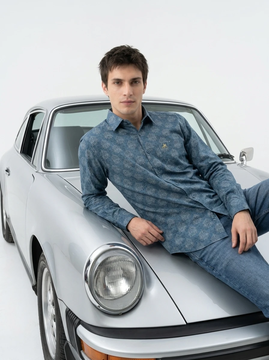 Man wearing a blue long-sleeve patterned button-down shirt and blue jeans, posing on the hood of a silver sports car.