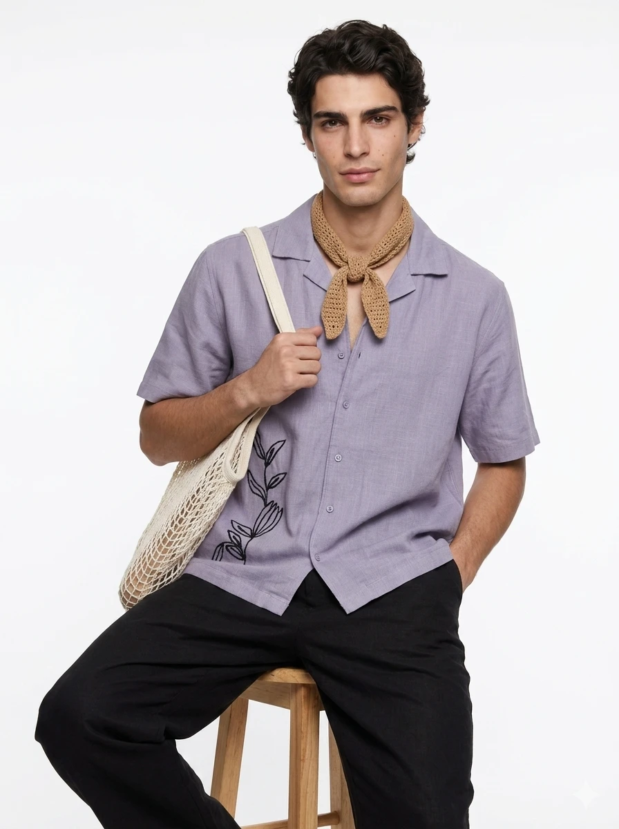 Man wearing a lavender short-sleeved button-up shirt with floral embroidery, a tan knit neck scarf, and a cream tote bag.