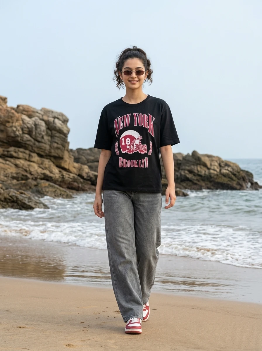 Woman walking on a sandy beach wearing a black graphic t-shirt and grey jeans.