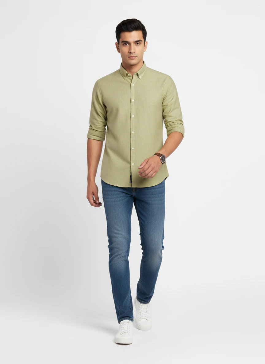 A male model wearing a light olive green long-sleeve button-down shirt and blue jeans against a plain white studio backdrop.