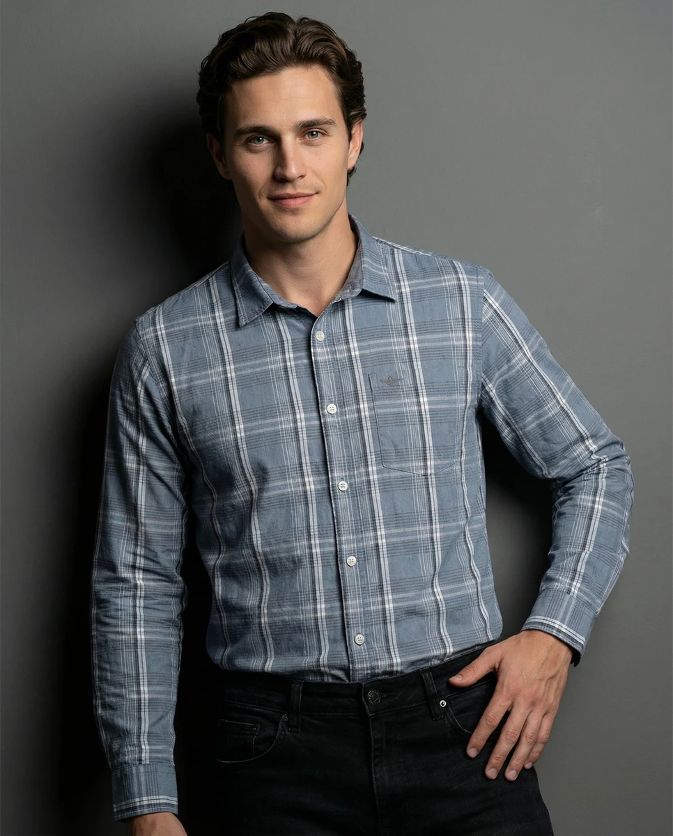 A man wearing a blue and white plaid long sleeve shirt and dark jeans against a neutral grey studio backdrop.