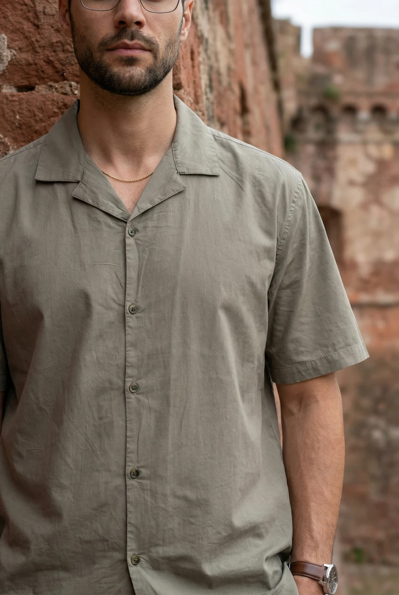 Close-up of a man wearing a short-sleeve olive green camp collar button-down shirt, standing against a textured brick wall.