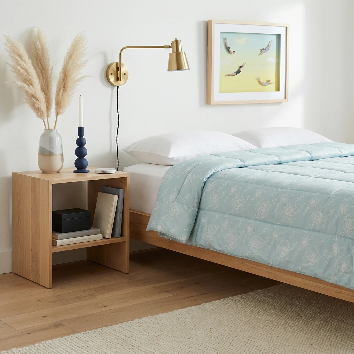Kanika comforter with light blue floral pattern draped on a bed next to a wooden nightstand in a bright bedroom.