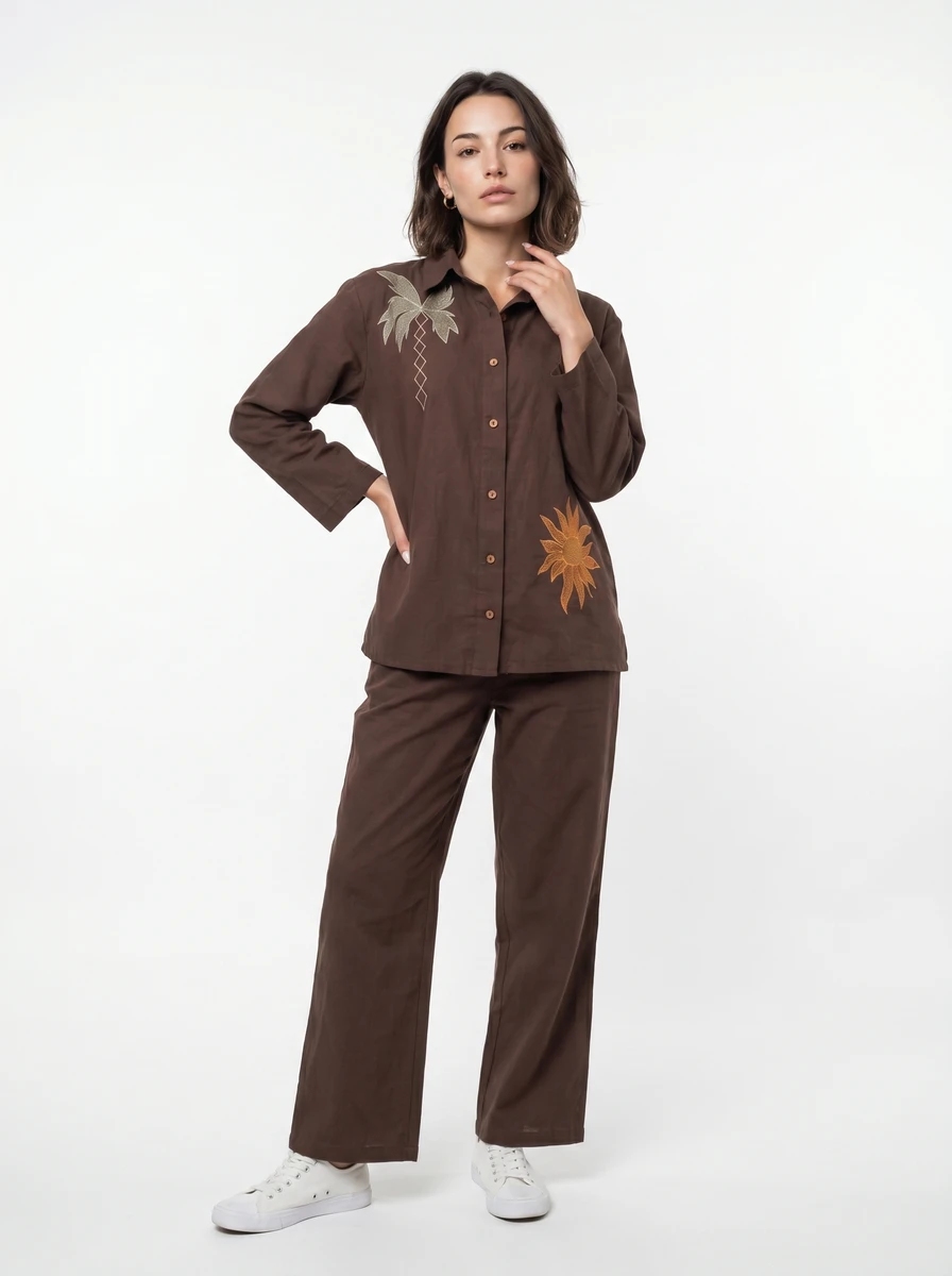 Model standing in a brown button-down shirt and pants set with embroidered palm and sun motifs, wearing white sneakers.