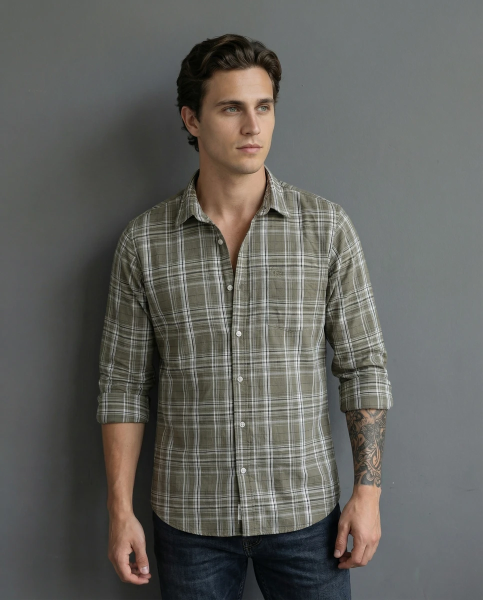 Man wearing an olive green plaid long-sleeve shirt and dark jeans against a grey studio wall.