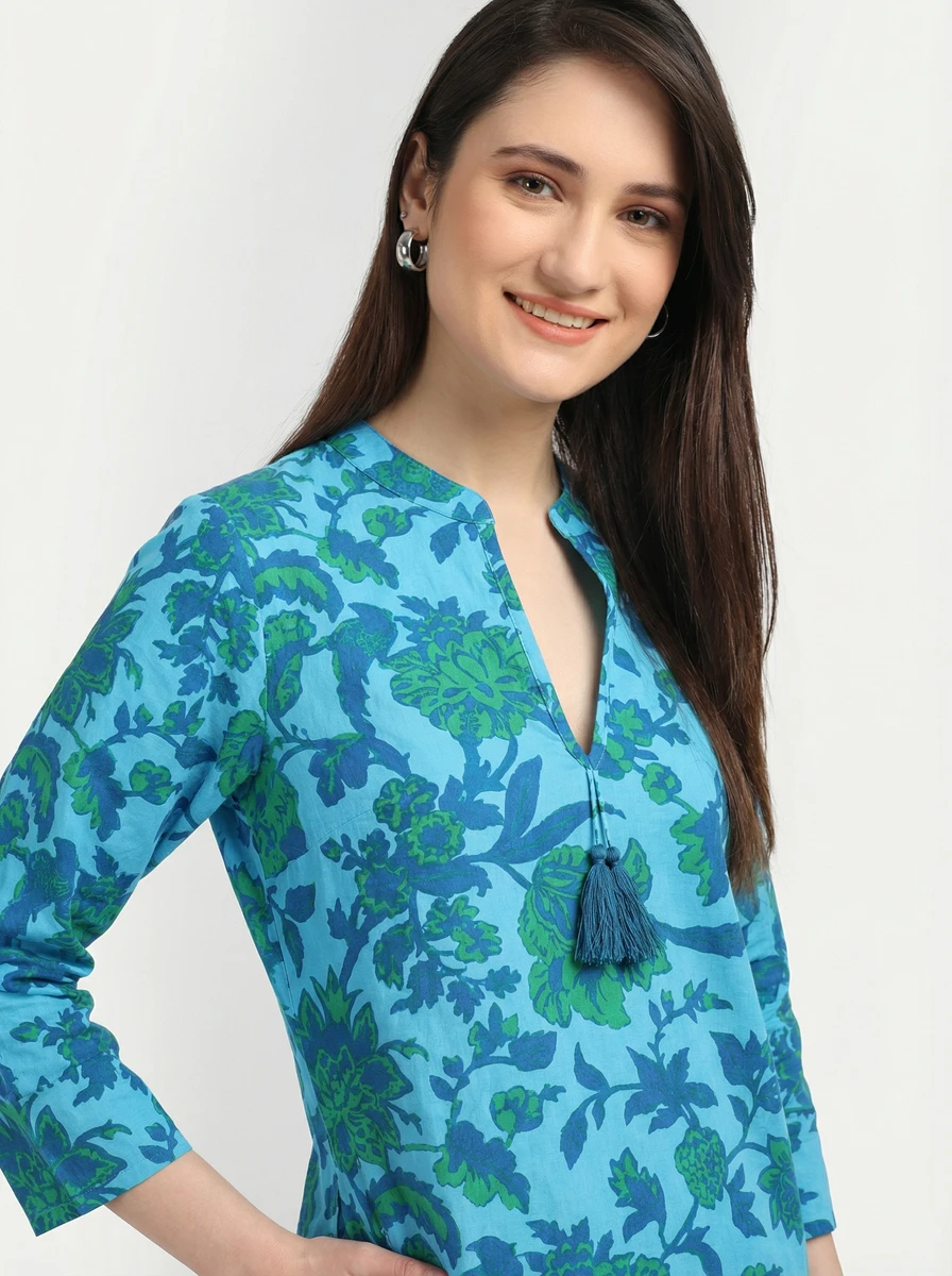 Woman wearing a bright blue tunic with a green floral print, v-neckline, and tassel ties.
