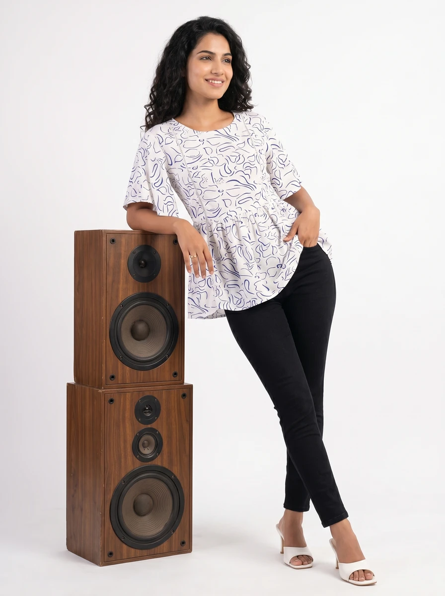 Woman wearing a white printed short-sleeve top and black trousers, leaning against wooden speakers.