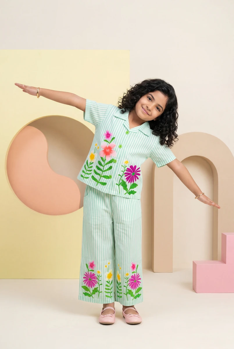 Young girl modeling a green and white striped kids pajama set with vibrant floral embroidery against a pastel studio backdrop.