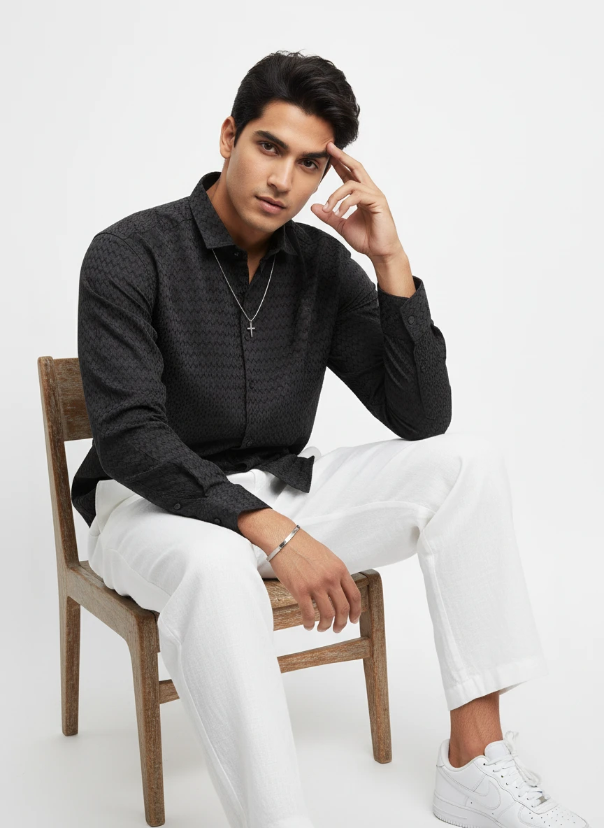 Man wearing a black patterned long-sleeve shirt and white trousers, seated on a wooden chair.