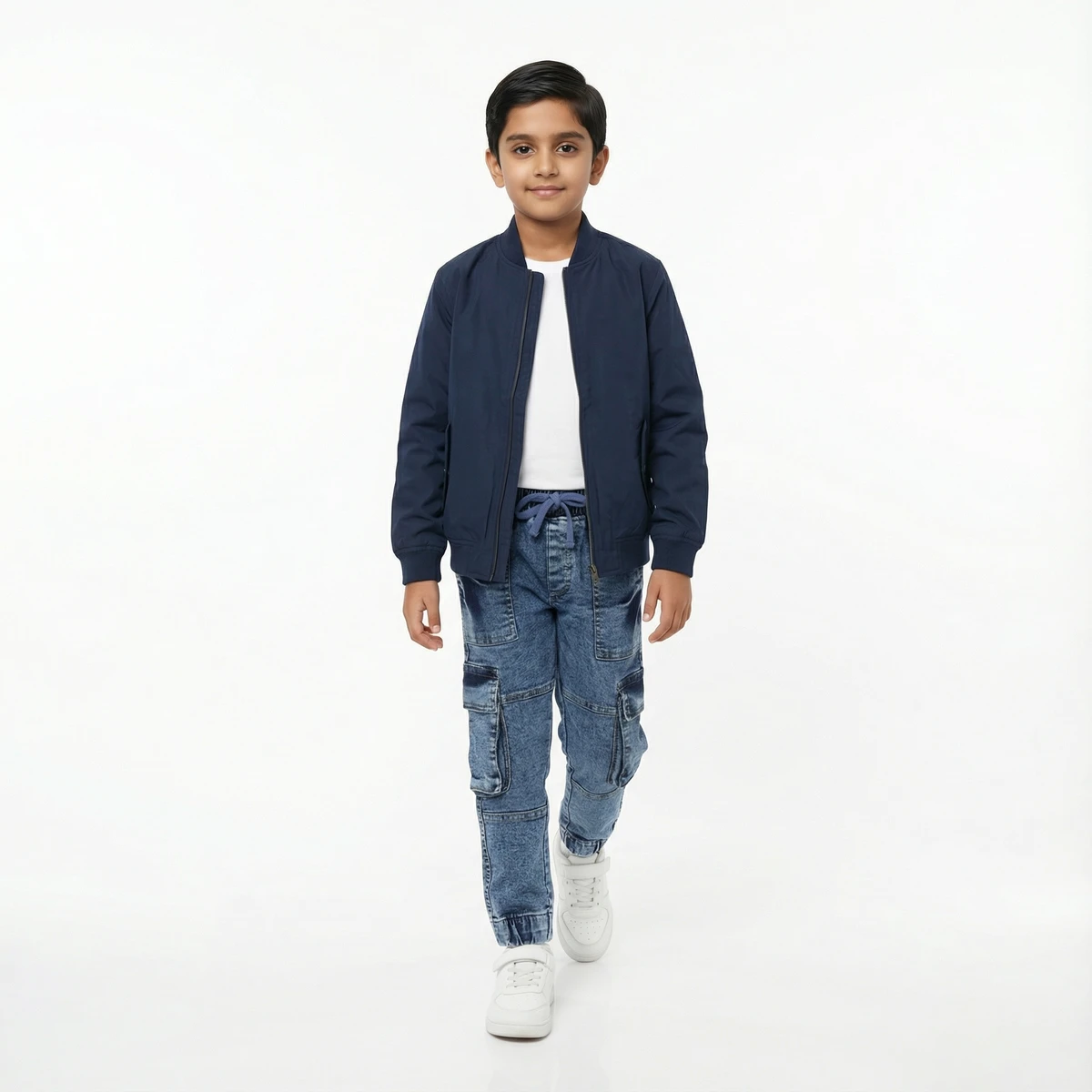 Young boy walking in a navy blue bomber jacket, white t-shirt, and blue denim cargo pants against a plain white background.