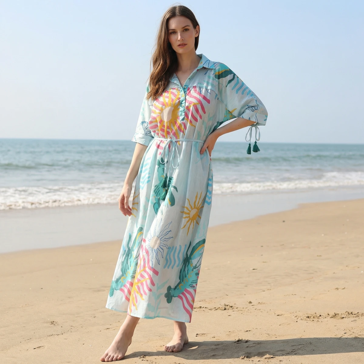 Woman wearing a light blue midi dress with colorful sun motifs posing on a sandy beach by the ocean.