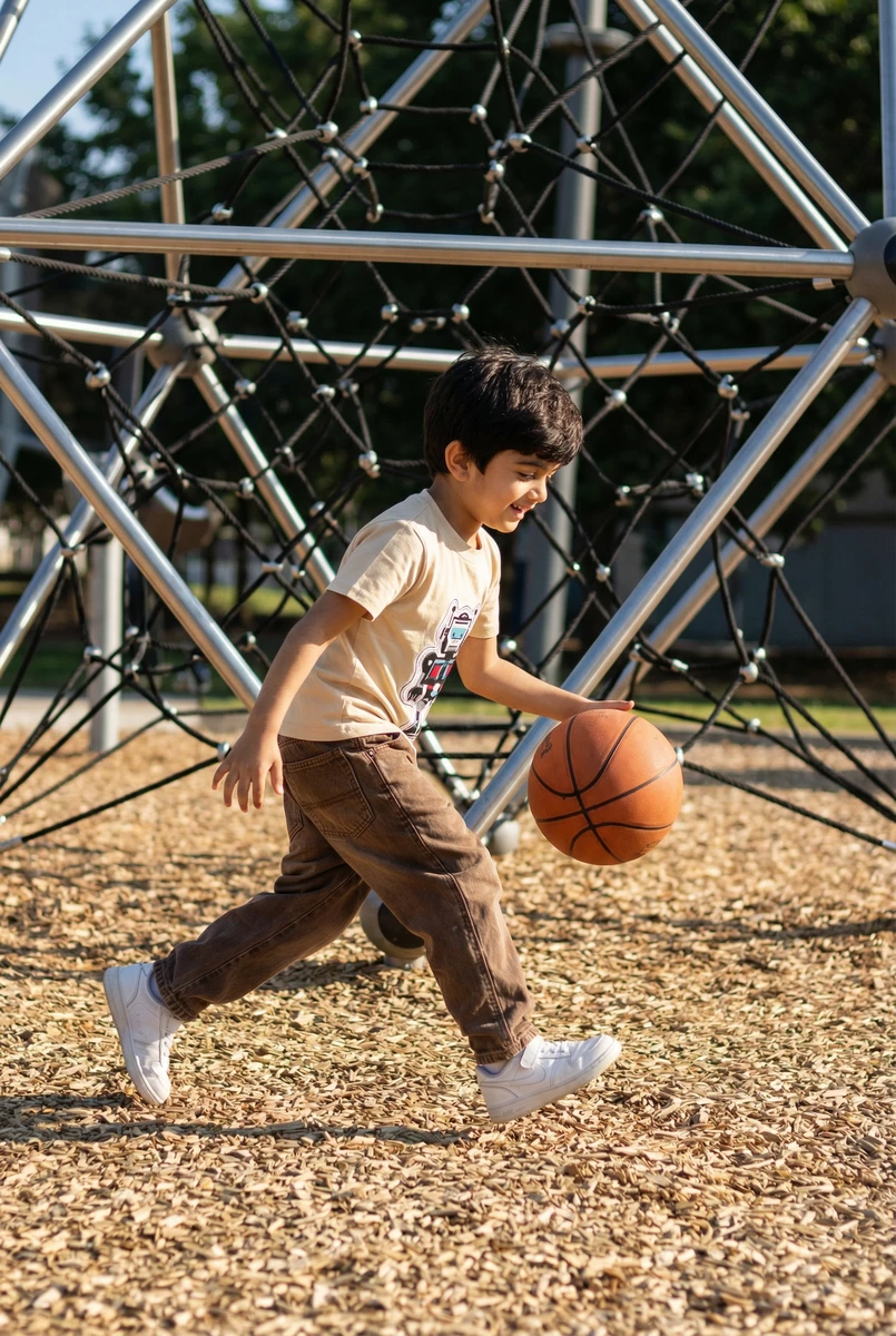 Toddler running at a playground wearing a beige graphic t-shirt and brown pants while bouncing a basketball.