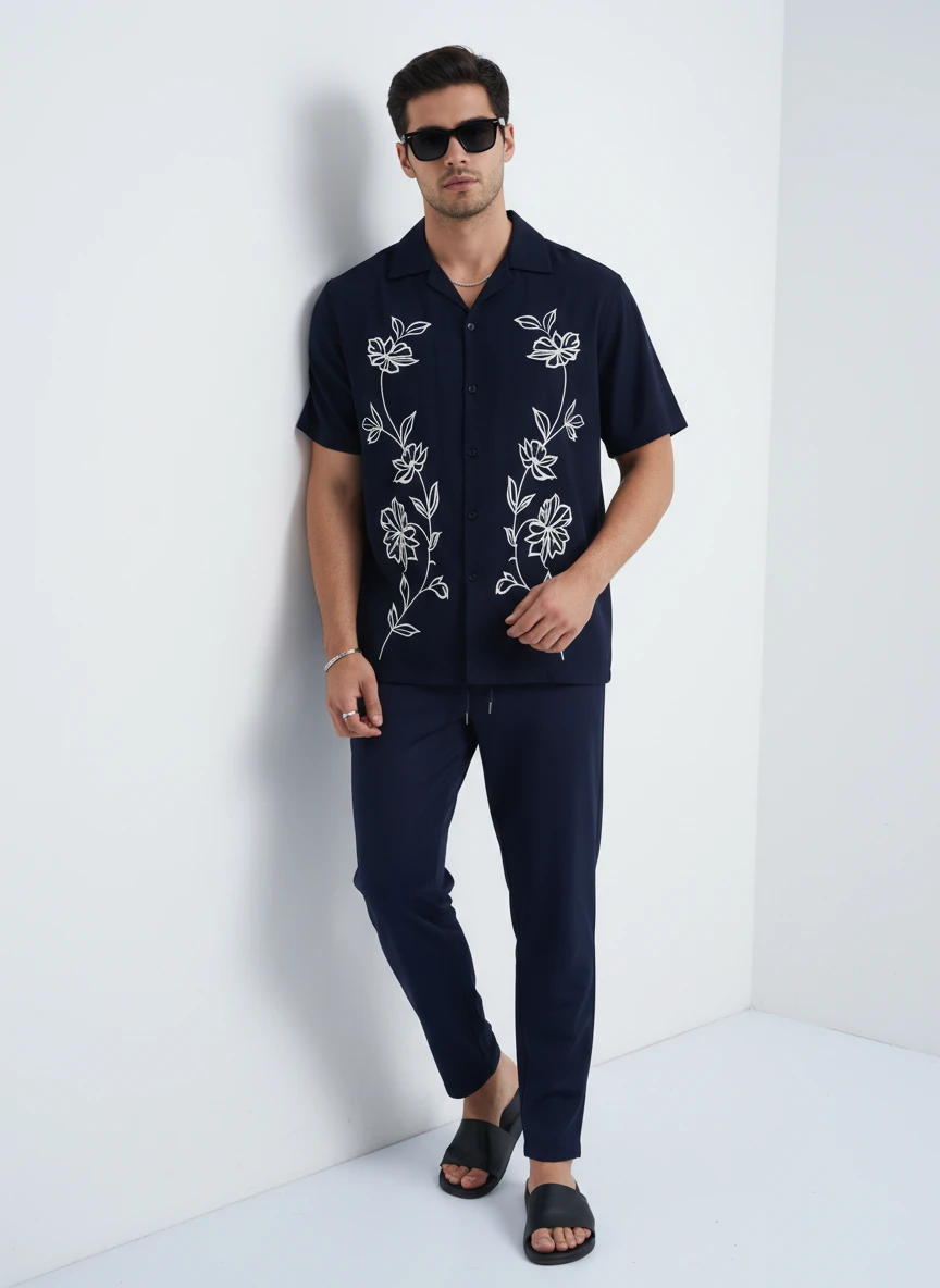 Man in a navy blue button-down shirt with white floral embroidery and matching trousers leaning against a white studio wall.