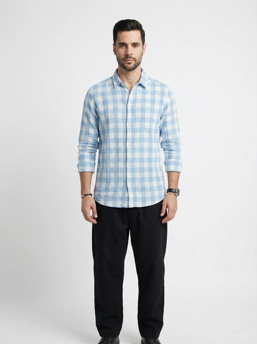 Man standing in studio wearing a light blue and white checkered long-sleeve shirt and black trousers.