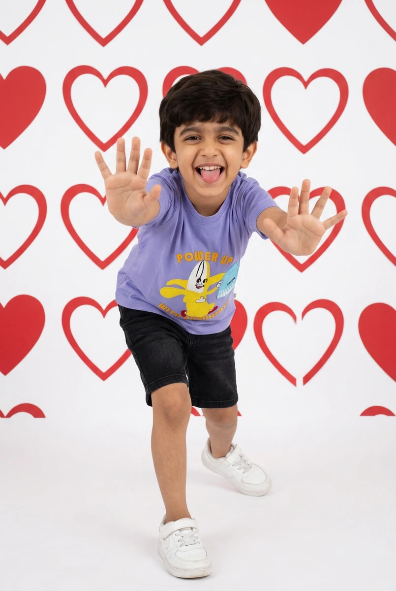 Toddler in a purple graphic t-shirt and black shorts poses with outstretched hands in front of a red heart patterned backdrop.